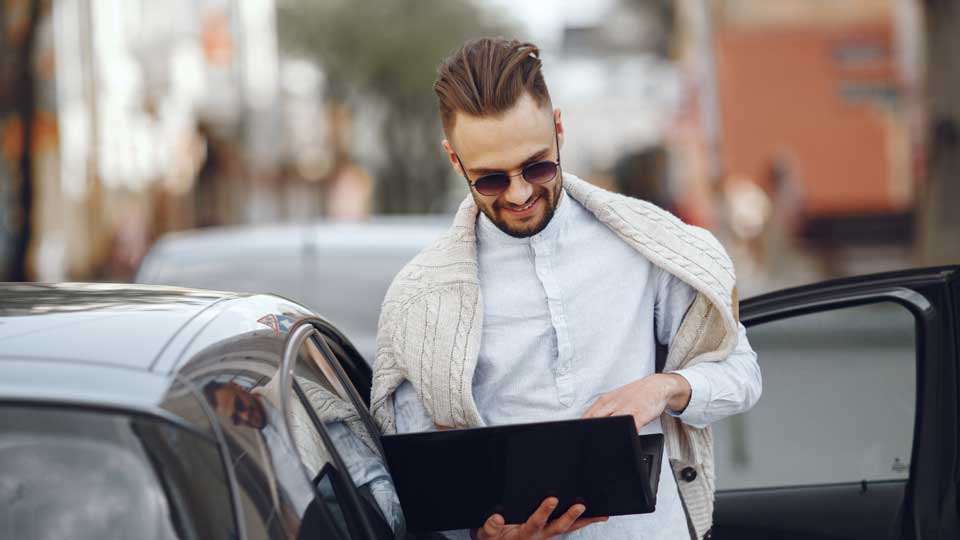 Mann steht neben einem Auto und schaut auf ein Tablet in der Hand